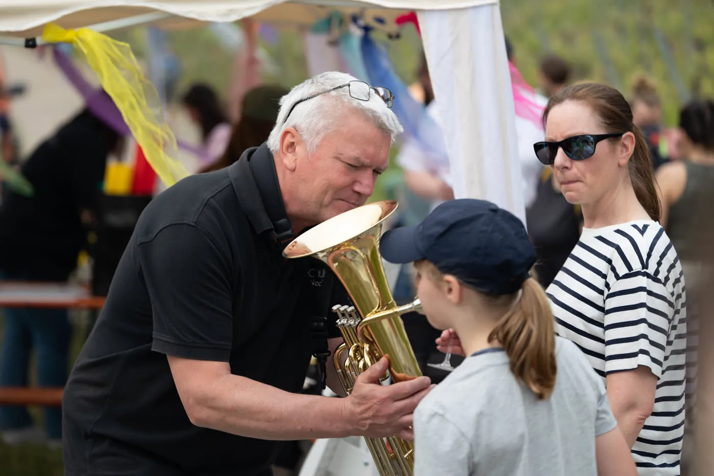 Jemand aus dem Team der Musikschule hält einem Mädchen das Euphonium, sie bläst zum ersten Mal hinein